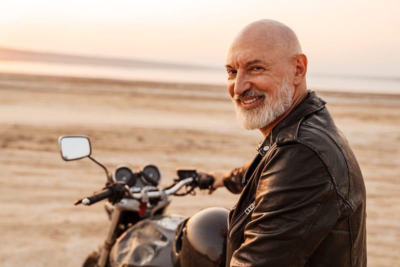 Mature bold man in leather jacket smiling while posing on motorbike